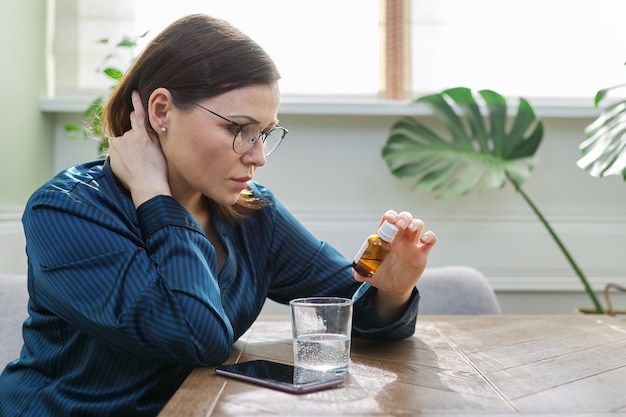 Mulher madura triste séria com remédio, copo de água, telefone. Mulher de pijama, sentada à mesa em casa, bebendo sedativos. Físico, psicológico, saúde mental, pessoas de meia-idade, copie o espaço