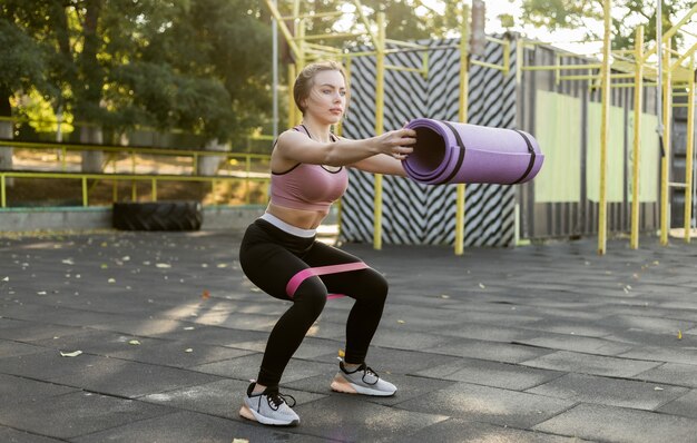 Mulher magra e apta exercita as pernas com elásticos de fitness e segurando o colchonete