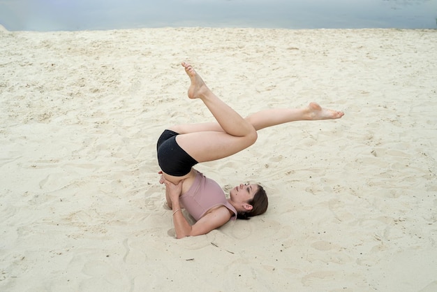 Mulher magra em roupas esportivas praticando ioga na praia de areia perto do lago no verão