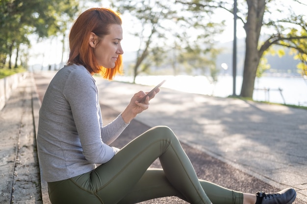 Mulher magro de gengibre em roupas de esporte, usando o telefone celular no parque. Saúde, bem-estar, estilo de vida fitness.