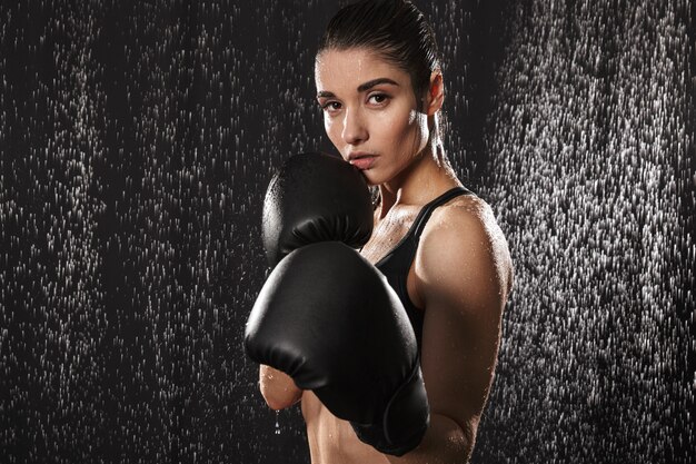 Mulher magro forte boxe usando luvas e em pé na posição de defesa sob gotas de chuva, isoladas sobre fundo preto