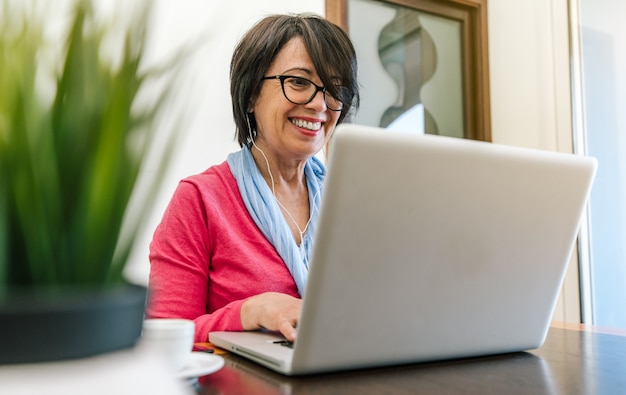 Mulher mais idosa sênior que trabalha com o laptop na mesa em casa interna. Pessoas maduras e conceito de tecnologia