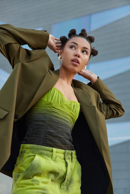 mulher mantém as mãos atrás da cabeça vestida com uma elegante jaqueta verde e jeans maquiagem brilhante posa contra um fundo cinza, pronta para passar o tempo com os amigos