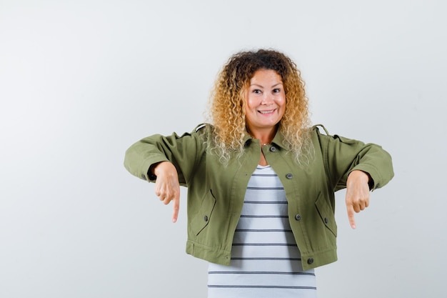 Mulher maravilhosa apontando para baixo enquanto sorria com jaqueta verde, camisa e olhando alegre, vista frontal.