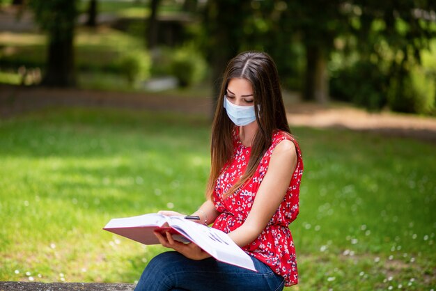 Mulher mascarada sentado em um parque e lendo um livro