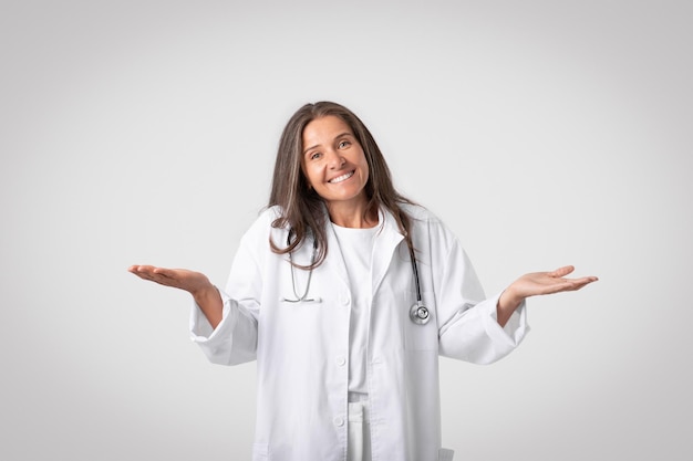 Mulher médica com estetoscópio tendo dúvidas espalhando as mãos de lado e levantando os ombros sorrindo para a câmera