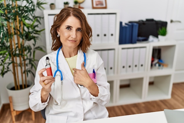 Mulher médica de meia-idade segurando cigarro eletrônico na clínica médica apontando o dedo para si mesma sorrindo feliz e orgulhosa