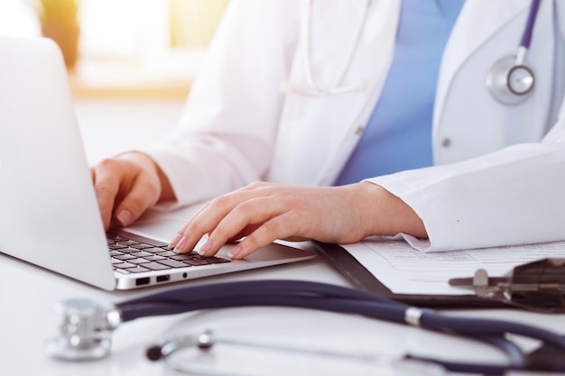 Mulher-médica desconhecida digitando no computador portátil enquanto está sentado à mesa na clínica ensolarada.