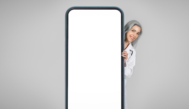 Mulher médica feliz espreitando por trás do grande fundo cinza da tela do smartphone