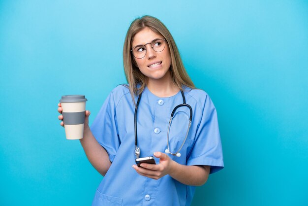 Mulher médica jovem cirurgião isolada em fundo azul segurando café para levar e um celular enquanto pensa em algo