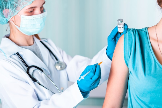 Mulher médica ou enfermeira de uniforme e luvas usando máscara protetora facial em laboratório, fazendo uma injeção segurando frasco de vacina com rótulo de vacina de Coronovírus COVID-19