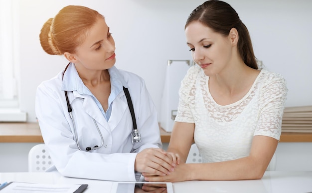 Mulher-médica tranquilizando sua paciente do sexo feminino enquanto está sentado na mesa. Conceito de medicina.