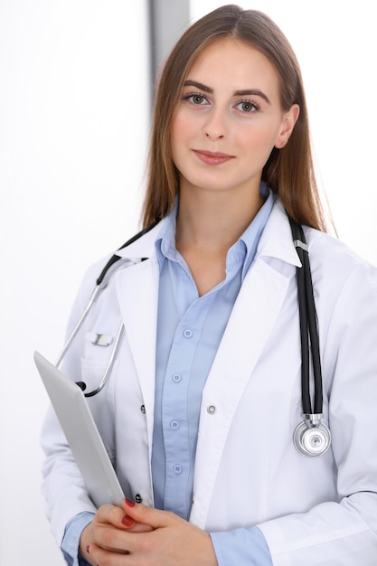 Mulher médica usando computador tablet em pé perto da janela no hospital. Médico feliz no trabalho. Conceito de medicina e cuidados de saúde.