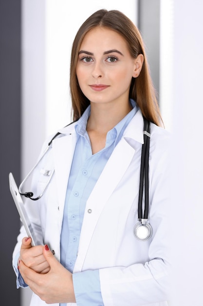 Mulher médica usando computador tablet em pé perto da janela no hospital. Médico feliz no trabalho. Conceito de medicina e cuidados de saúde.