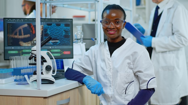 Mulher médico negro sorrindo para a câmera, sentado no moderno laboratório científico equipado. Equipe multiétnica examinando a evolução do vírus usando ferramentas químicas de alta tecnologia para pesquisa científica e desenvolvimento de vacinas