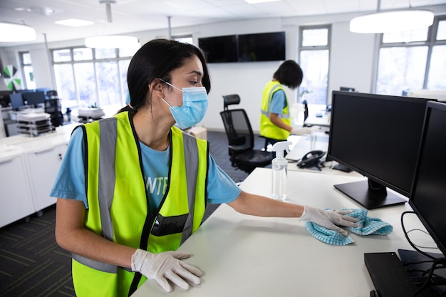 Foto mulher mestiça e colega usando coletes de alta visibilidade, luvas e máscaras faciais higienizando um escritório com desinfetante. higiene no local de trabalho durante a pandemia do coronavirus covid 19.