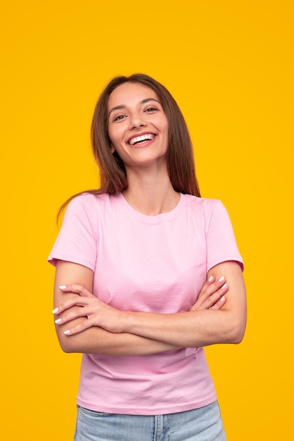 Mulher milenar alegre em roupa casual com os braços cruzados, olhando para a câmera e rindo alegremente contra um fundo amarelo brilhante