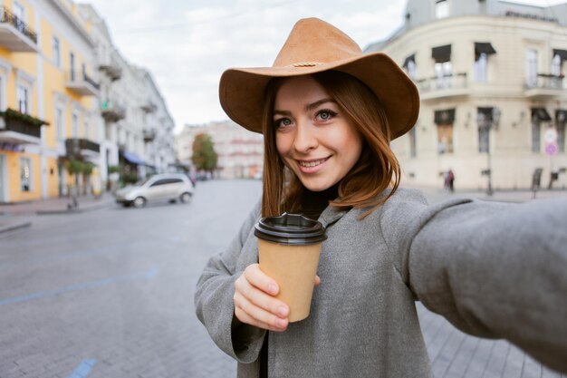 Mulher milenar elegante em um casaco mais liso e chapéu de feltro sacode uma xícara de café em movimento e faz selfie na cidade europeia