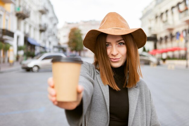 Mulher milenar elegante em um casaco mais liso e chapéu de feltro sacode uma xícara de café em movimento enquanto caminha em uma cidade europeia