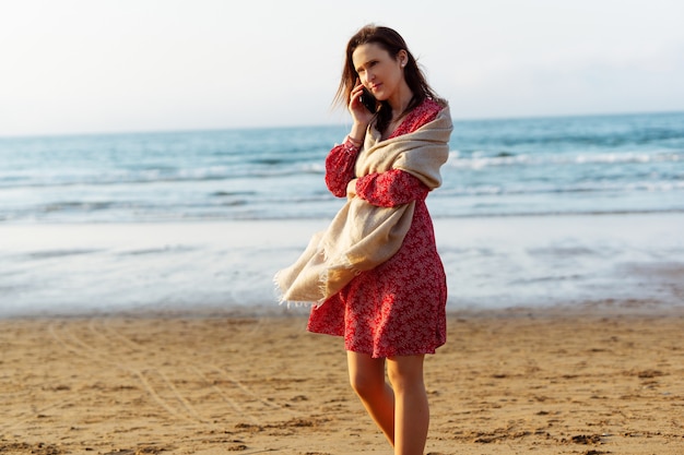mulher milenar falando no smartphone na praia com vestido vermelho