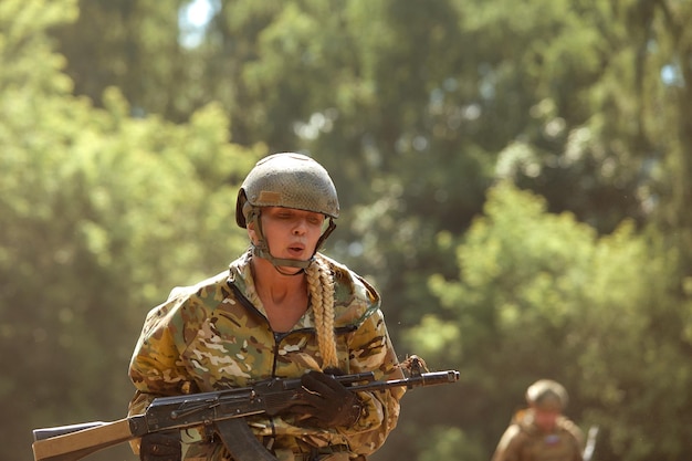 Mulher militar caucasiana em equipamento tático posando para foto na temporada de verão vestindo uniforme camuflado verde e rifle de assalto em equipamento militar e fone de ouvido está olhando para o lado