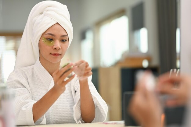 Foto mulher millennial atraente aplicando anti-fatiga sob os olhos refletindo-se no espelho da mesa de roupa