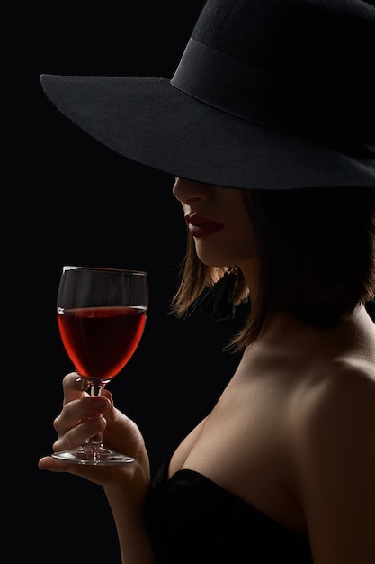 Mulher misteriosa elegante de chapéu segurando um copo de vinho tinto