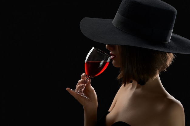 Mulher misteriosa elegante de chapéu segurando um copo de vinho tinto