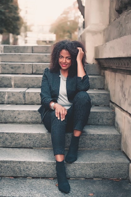 Mulher misturada nova com penteado afro que sorri no fundo urbano. Garota negra, vestindo roupas casuais.