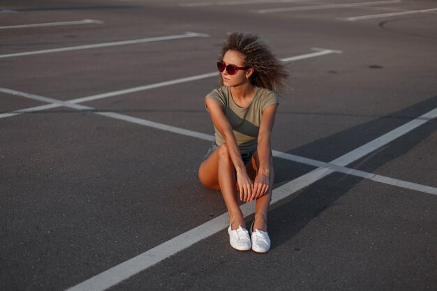 Mulher modelo elegante e bonita jovem hippie com penteado encaracolado com óculos de proteção solar em shorts jeans com uma camiseta elegante e tênis branco sentado no asfalto na rua