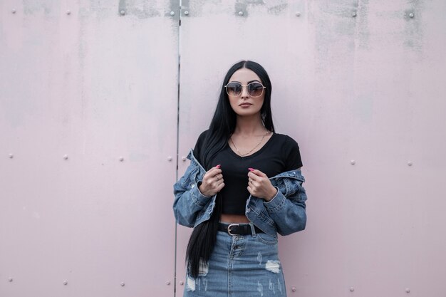 Mulher moderna bonita jovem hippie em elegantes óculos de sol pretos levanta e endireita uma jaqueta jeans perto de uma parede vintage. modelo de menina morena atraente com roupas da moda, posando ao ar livre.