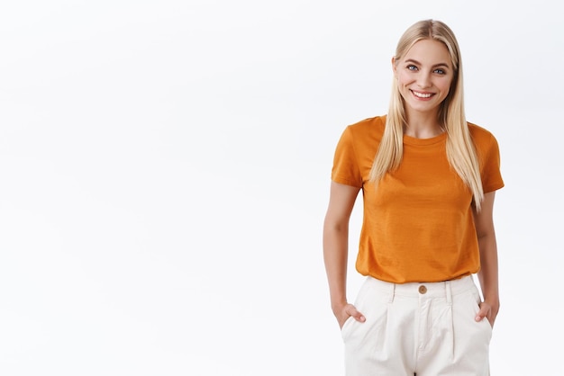 Mulher moderna elegante e atraente com cabelo loiro usa camiseta laranja de mãos dadas nos bolsos das calças sorrindo alegre e entretido parece confiante e despreocupado em pé fundo branco