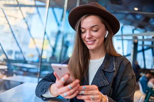 Mulher moderna elegante elegante hipster de chapéu ouve música on-line usando telefone celular e fones de ouvido sem fio em um café. Estilo de vida digital