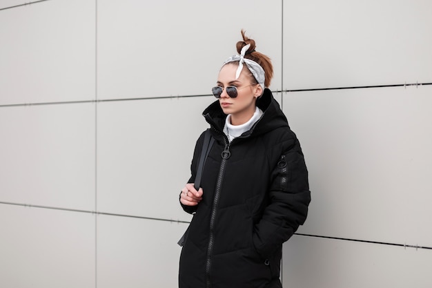 Mulher moderna jovem hippie em óculos de sol redondos com um casaco elegante em uma bandana da moda com uma mochila de couro está de pé perto de uma parede branca num dia quente de inverno. Garota americana elegante de férias