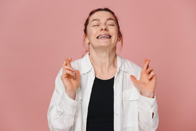 Mulher moderna sorridente com aparelhos ortopédicos cruzando os dedos em um fundo rosa