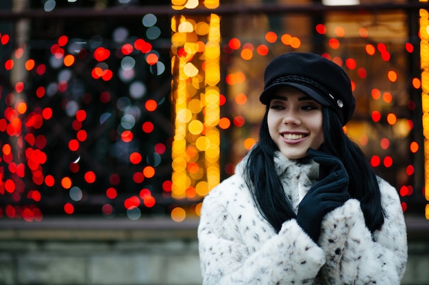 Mulher morena alegre com boné preto posando na rua sobre um fundo de guirlandas com bokeh