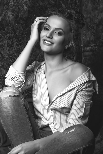 Mulher morena alegre com maquiagem natural usa camisa e calça jeans posando no estúdio. Retrato preto e branco
