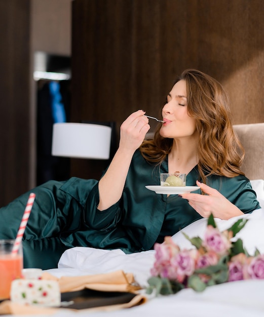 Mulher morena alegre, desfrutando de comer sobremesa na cama enquanto descansa no quarto de hotel Senhora atraente de pijama elegante tendo um dia de folga relaxando sozinho Estilo de vida luxuoso Jovem glamourosa