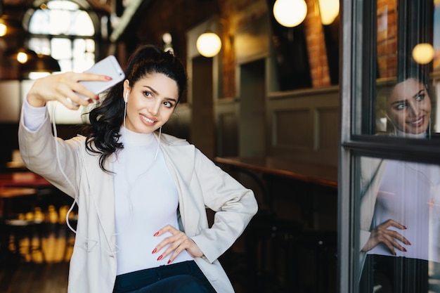 Mulher morena alegre em roupas elegantes brancas segurando o telefone celular genérico fazendo sefie contra o fundo do café aconchegante elegante jovem fêmea fazendo foto de si mesma se divertindo Tecnologia moderna