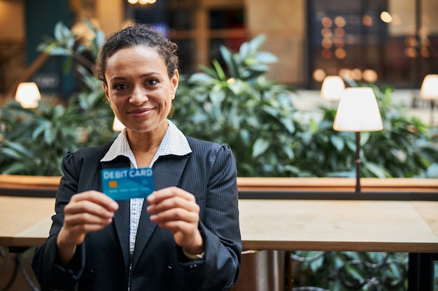 Mulher morena alegre segurando seu cartão de débito com as duas mãos enquanto recomenda serviços bancários online