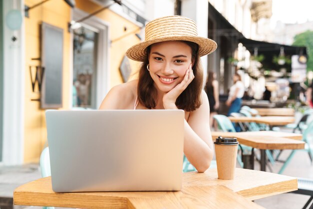 mulher morena alegre sorrindo e usando o laptop enquanto está sentada na rua, no café de verão, tomando um café para viagem