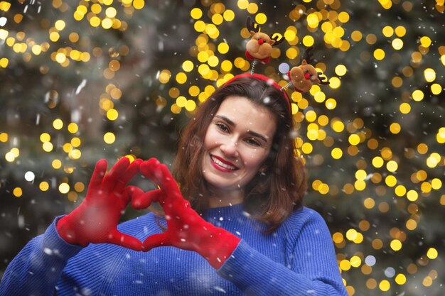 Mulher morena alegre usa aro de cabelo engraçado, mostrando o sinal de coração durante a queda de neve. Espaço vazio