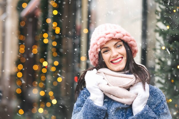 Mulher morena alegre usa boné rosa claro de malha e lenço andando na cidade decorada com guirlandas durante a queda de neve. Espaço para texto