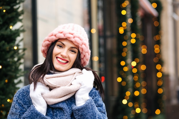 Mulher morena alegre usa boné rosa claro de malha e lenço andando pela cidade decorada com guirlandas. Espaço para texto