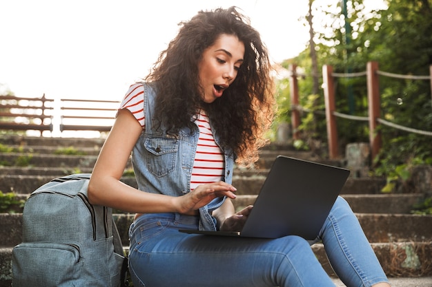 Mulher morena animada sentada na escada do parque num dia de verão e usando laptop prata com surpresa