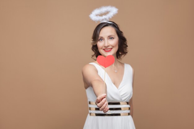 Mulher morena anjo de meia idade em um vestido branco com nimbo no topo da cabeça dando um adesivo de coração vermelho e olhando para a câmera com um sorriso. Foto de estúdio, interna, isolada em fundo marrom claro