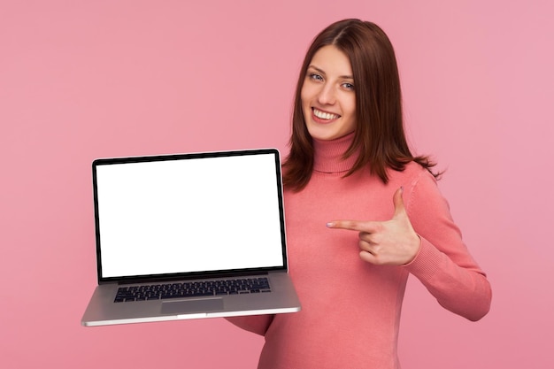 Mulher morena apontando o dedo para o laptop com tela em branco ela segurando na mão mostrando a tela vazia se gabando com anúncio na internet Tiro de estúdio interno isolado no fundo rosa