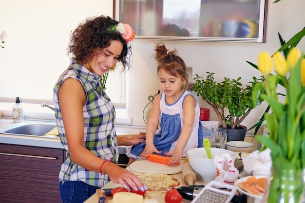 Mulher morena atraente com cabelos cacheados e sua filha pequena cozinhando comida em uma cozinha de casa.