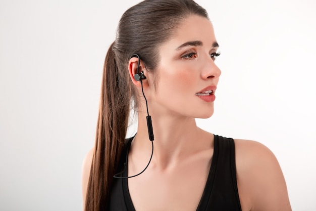 Foto mulher morena atraente com top preto para correr ouvindo música em fones de ouvido posando isolada em um penteado de rabo de cavalo de parede branca