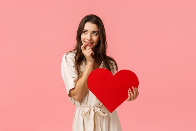 Mulher morena atraente e atraente, sonhadora, bonita e sedutora em vestido, olhar data perfeita de imagem esquerda, tocando o lábio e sorrindo com expressão tentadora e ansiosa, segurando o coração dia dos namorados, conceito de amor